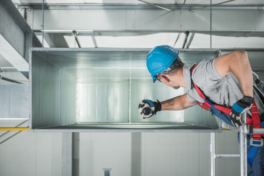 Commercial Warehouse Air Ventilation and Climate Control Canal Maintenance by Caucasian Technician in His 30s. Men Checking Inside Rectangle Canal Interior.